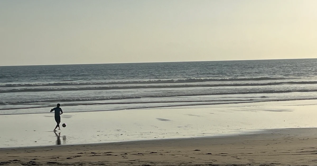 A person playing football alone on Candolim beach at dusk, with gentle waves and a hazy evening sky