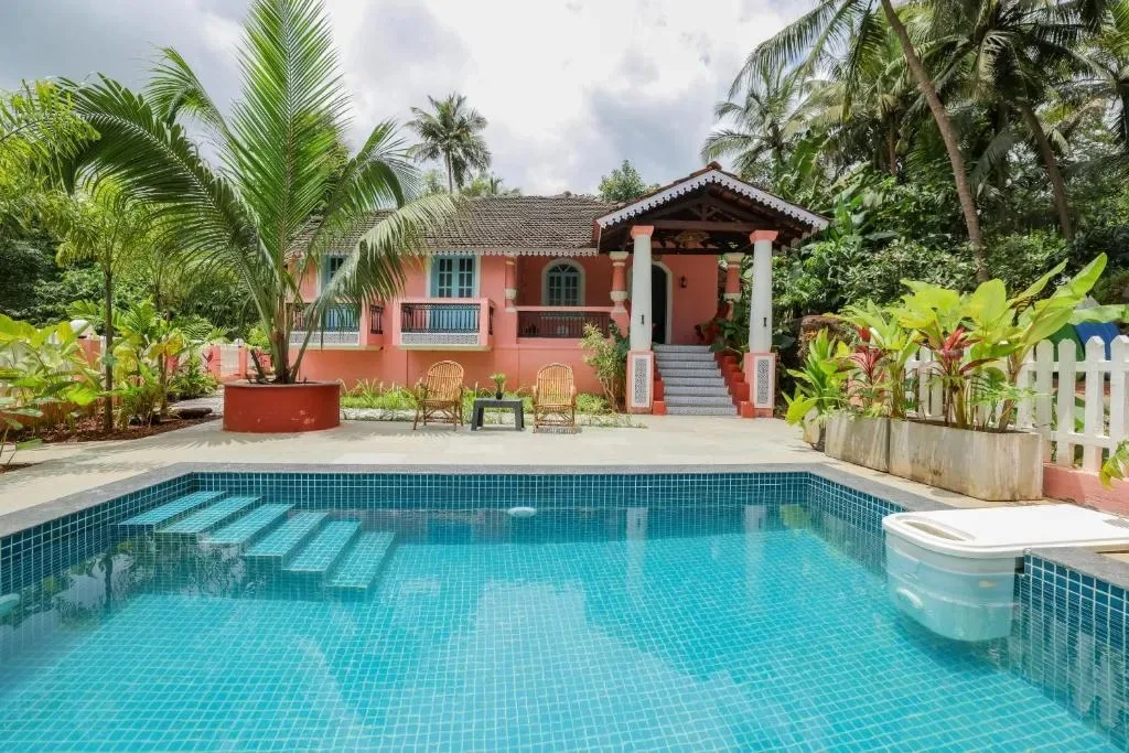 A cozy pink heritage homestay cottage in Goa with a clean swimming pool in the foreground surrounded by palms