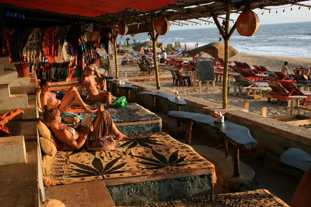 Rustic beach shack in Anjuna, Goa, featuring floor mattresses, cushions, and relaxed tourists reading and looking out at the ocean waves