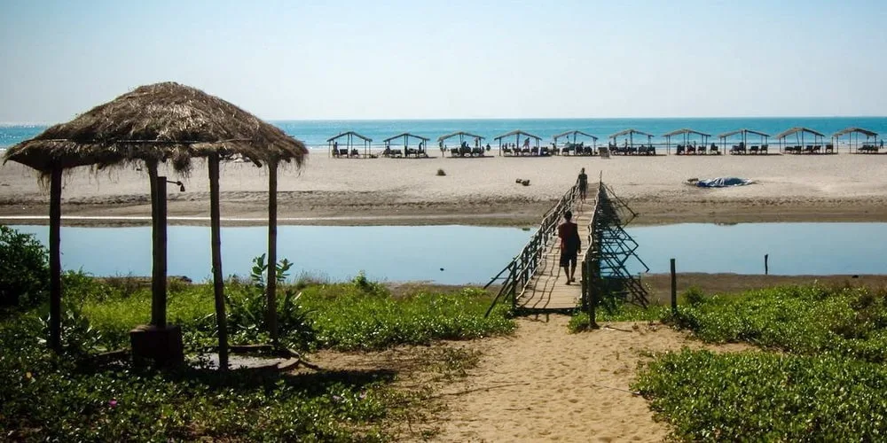Classic wooden footbridge over the Mandrem creek with a traveler walking towards the sandy beach and shacks in North Goa