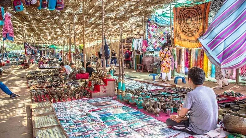 A vendor sitting amidst traditional brass vessels, singing bowls, and textiles at a flea market in Goa