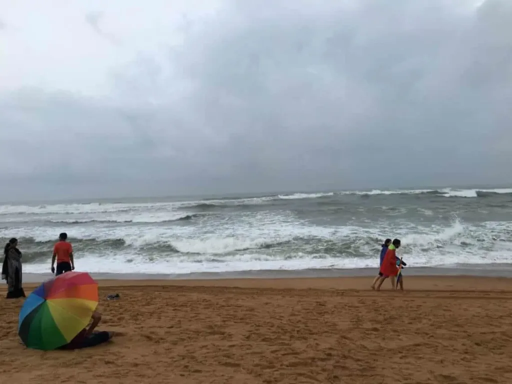 People walking on a wide beach with dark sand and big crashing monsoon waves under overcast skies