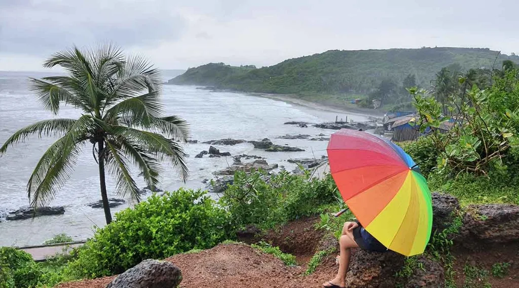 A traveler sitting on a cliff in Goa under a rainbow umbrella overlooking the stormy monsoon sea