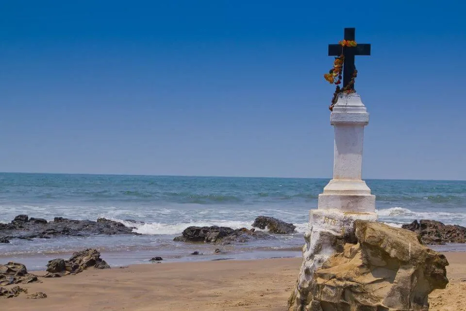 A white stone monument cross standing on a rocky outcropping on Morjim beach in front of the breaking waves and blue sea